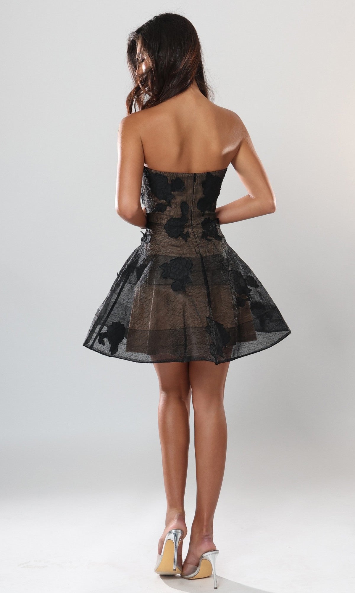 A woman stands facing away against a plain background, wearing the Jovani 53779 short homecoming dress— a strapless black design featuring a sheer floral lace overlay—paired with clear high-heeled shoes.